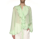 A person wears the Victoria Beckham Romantic Blouse—a sheer, light green top with blouson sleeves and ruffle details—paired with white pants, standing against a white background.