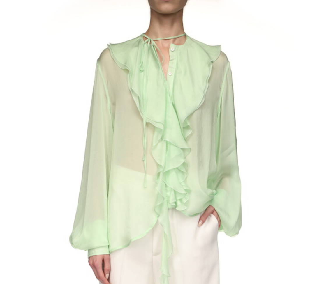 A person wears the Victoria Beckham Romantic Blouse—a sheer, light green top with blouson sleeves and ruffle details—paired with white pants, standing against a white background.