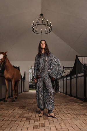 A woman in the Johanna Ortiz La Bohemia Top stands in a stable aisle with two horses, one brown and one white, under a black chandelier.