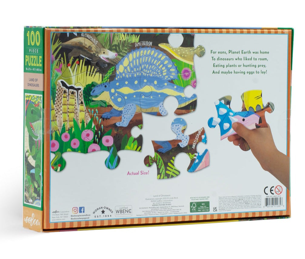 A person is holding a piece from the eeboo Land of Dinosaurs 100 Piece Puzzle by Eeboo next to its box, which captivates paleontology enthusiasts with its depiction of dinosaurs in their natural habitat and intriguing information about their ancient existence.