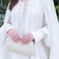 A person wearing the Lanvin Long Sleeve Blouse Open Neck, adorned with a delicate lace-up neckline, holds a small white beaded clutch in both hands against a backdrop of lush green foliage.