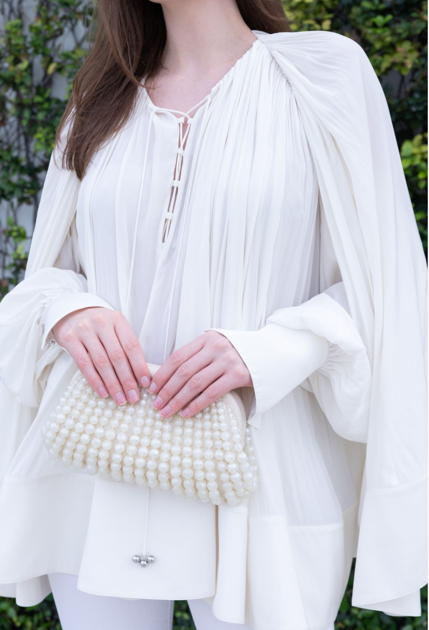 A person wearing the Lanvin Long Sleeve Blouse Open Neck, adorned with a delicate lace-up neckline, holds a small white beaded clutch in both hands against a backdrop of lush green foliage.