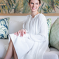 A woman in a Lanvin Flared Pleated Dress with Long Sleeves sits on a sofa with green and blue patterned cushions and a leafy backdrop, smiling and looking to the side.