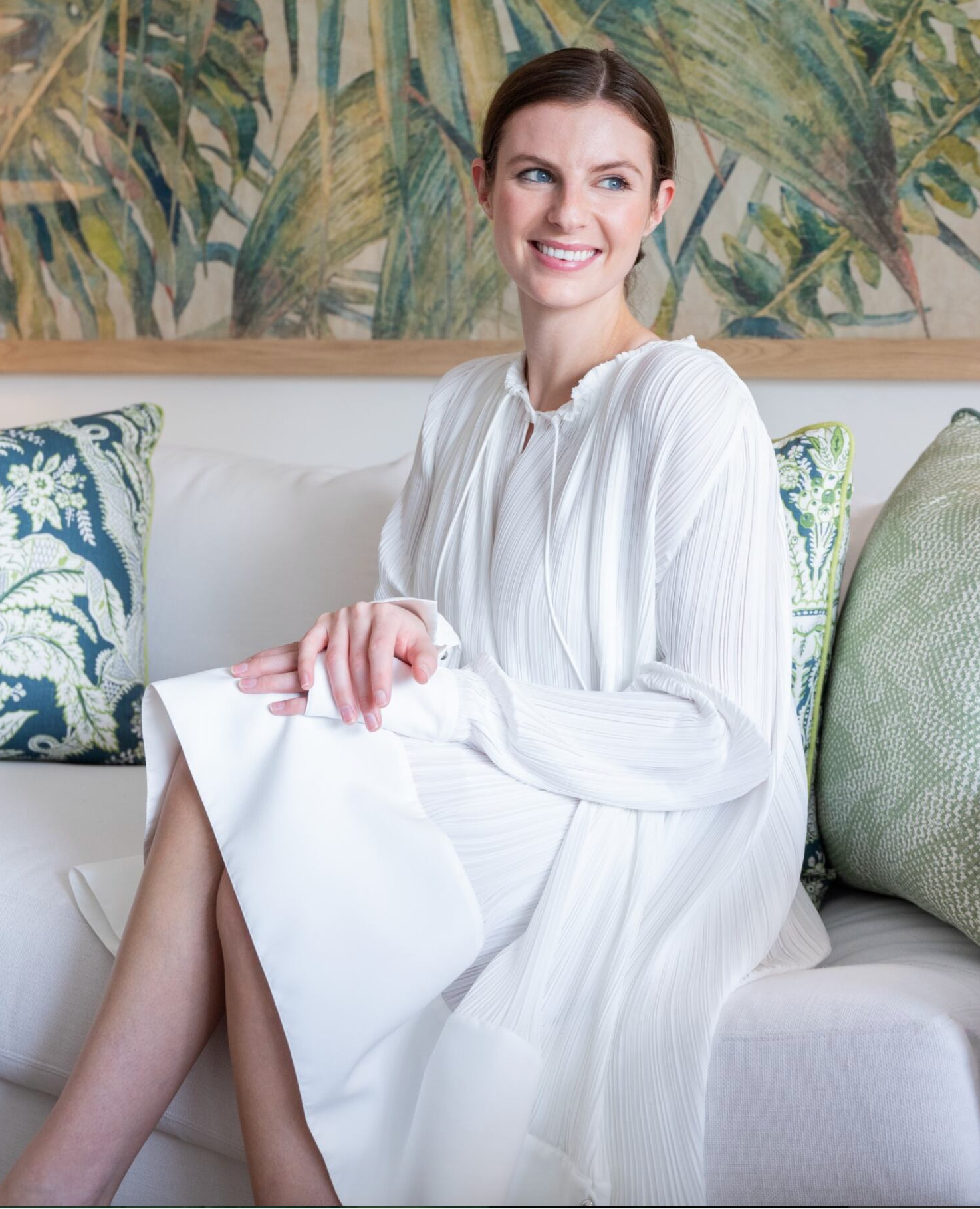 A woman in a Lanvin Flared Pleated Dress with Long Sleeves sits on a sofa with green and blue patterned cushions and a leafy backdrop, smiling and looking to the side.