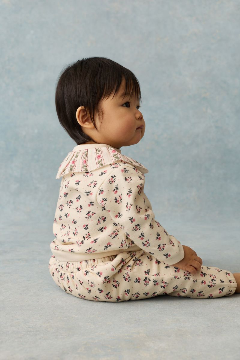 A baby sits on the floor in profile against a light blue background, wearing the Louise Misha Baby Girls’ Rosaline Sweatshirt—a cream-colored floral top featuring a ruffle neckline and decorative collar.