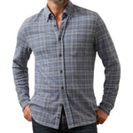 A man wearing a Faherty Legend Sweater Shirt in blue and gray plaid with long sleeves and dark jeans stands with one hand in his pocket.