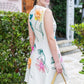 A woman in a white Lela Rose Embroidered Canvas Mini Dress, adorned with colorful floral embellishments, holds a yellow handbag as she stands on a stone path in front of lush greenery and a charming building.