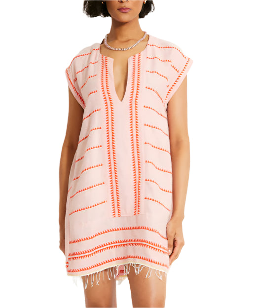 The individual wears the lemlem Elina Caftan by Lemlem, a light pink dress with orange patterned stripes and a fringed hem, crafted from hand-spun cotton.