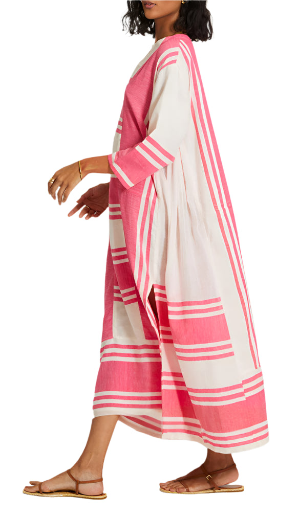 A woman models the Lemlem Fana Caftan—long, loose-fitting with pink and white stripes—paired with flat sandals and gold jewelry, walking sideways against a white backdrop for a look that's effortlessly versatile.