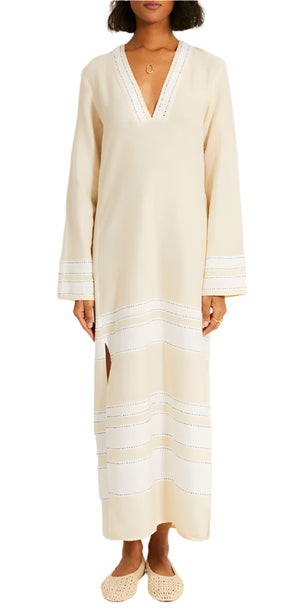 Wearing the Lemlem Theodora Column Dress, a bohemian beige dress with white stitched stripes, side slits, long sleeves, and a V-neck. They complete the outfit with beige slip-on shoes.