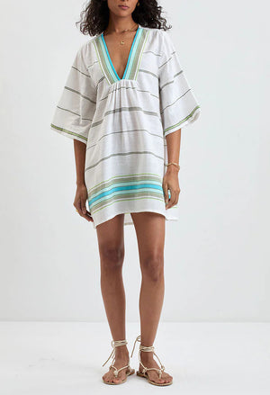 A woman stands against a plain background wearing the Lemlem Belkis Caftan—white, hand-woven cotton with green and blue stripes, a V-neckline, wide sleeves—and sandals.