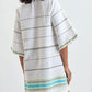 A woman faces away, wearing the Lemlem Belkis Caftan—a loose, knee-length bohemian dress in hand-woven cotton with horizontal stripes and green and blue accents near the hem.