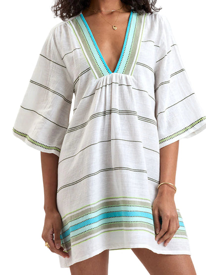 A person wearing the Lemlem Belkis Caftan, a white hand-woven cotton dress with green and blue stripes and wide sleeves, styled with gold jewelry.