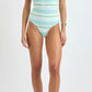 A woman stands against a plain background wearing the Lemlem Dinha One Piece, a high-cut, striped, ribbed swimsuit. She pairs it with brown sandals, a necklace, and a bracelet. Her arms rest relaxed at her sides.