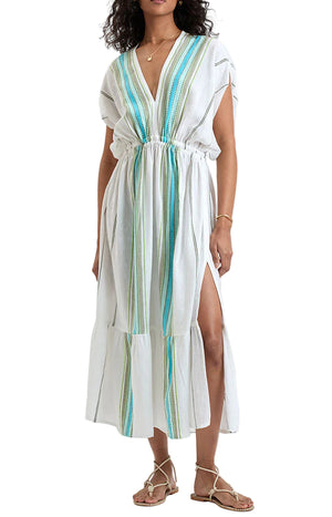 Wearing Lemlem's Leila Plunge Dress—a hand-woven cotton white midi with blue and green stripes, short sleeves, cinched waist, and side slit—paired with flat sandals for effortless resort style.