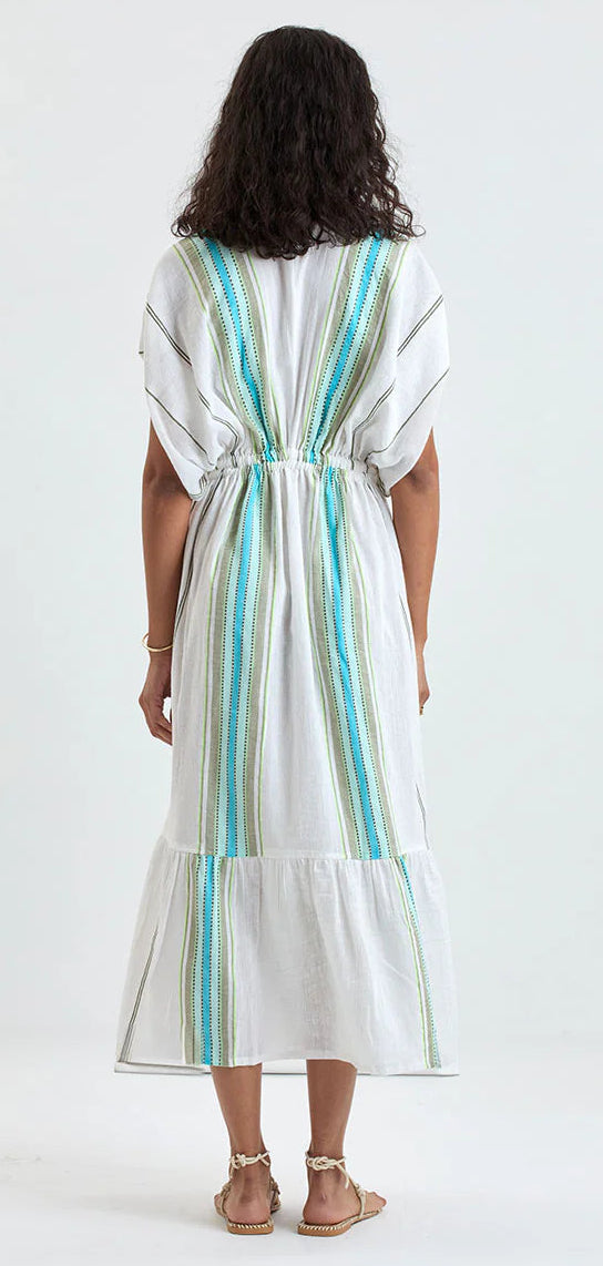 A person stands facing away, wearing the Lemlem Leila Plunge Dress—a hand-woven cotton piece with blue and green vertical stripes, a cinched waist, and short sleeves. Styled with sandals, this effortless dress is perfect for resort wear.