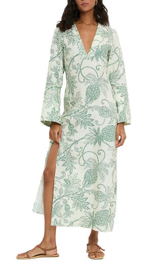 A woman wears the Lemlem Theodora Column Dress—long, light-colored European flax linen with green botanical patterns, long bell sleeves, deep V-neck, and a thigh-high slit—styled with flat sandals.