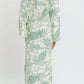 A person stands facing away, wearing Lemlem’s Theodora Column Dress—crafted from European flax linen with green botanical patterns—and sandals, set against a plain white background.