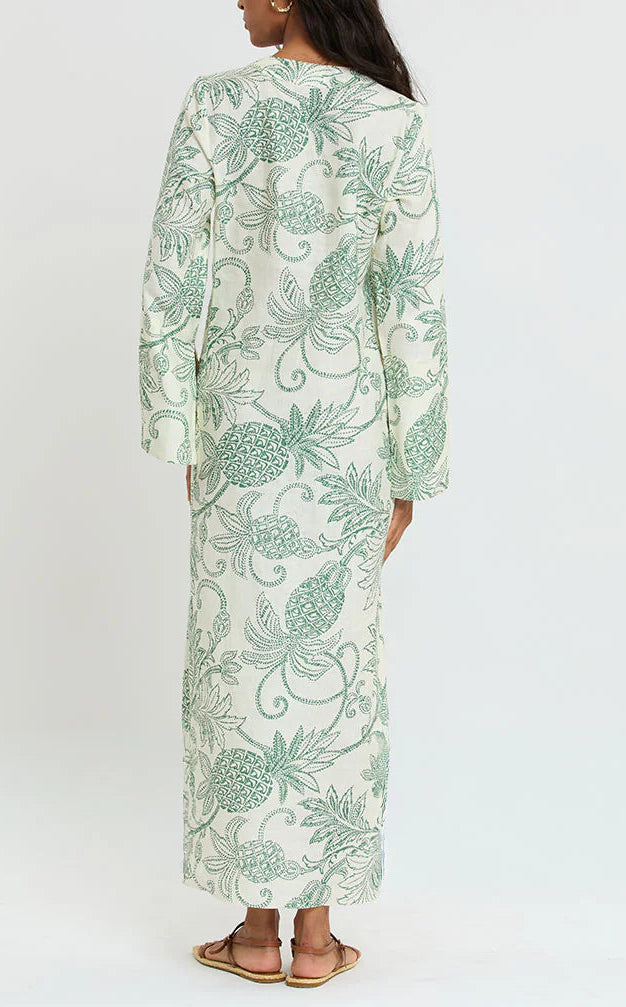 A person stands facing away, wearing Lemlem’s Theodora Column Dress—crafted from European flax linen with green botanical patterns—and sandals, set against a plain white background.