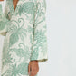 A person wearing Lemlem’s Theodora Column Dress in cream European flax linen, detailed with green embroidered pineapple and leaf motifs, long sleeves, and a deep V-neckline.