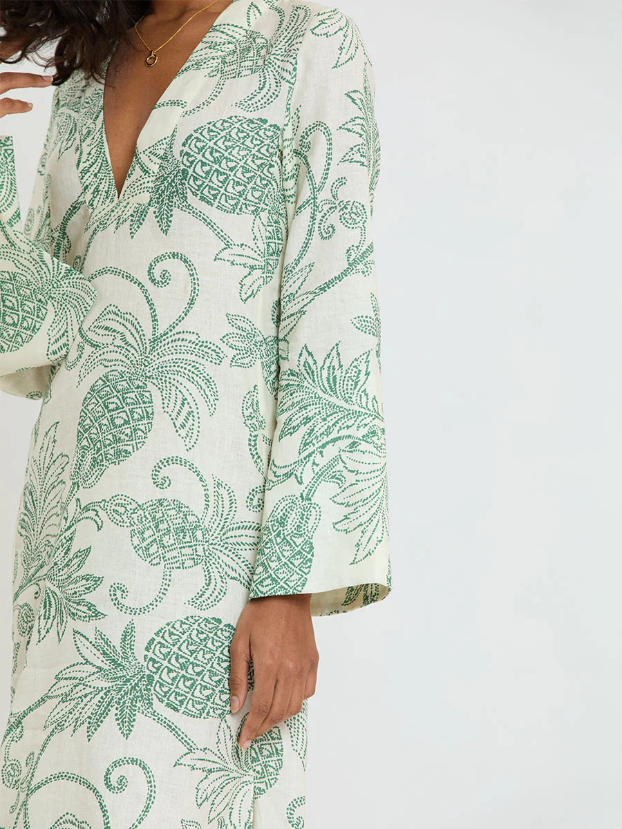 A person wearing Lemlem’s Theodora Column Dress in cream European flax linen, detailed with green embroidered pineapple and leaf motifs, long sleeves, and a deep V-neckline.
