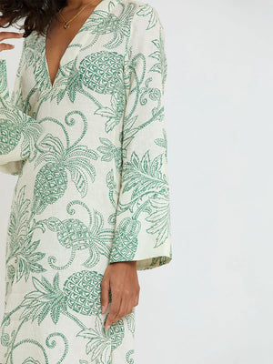 A person wearing Lemlem’s Theodora Column Dress in cream European flax linen, detailed with green embroidered pineapple and leaf motifs, long sleeves, and a deep V-neckline.