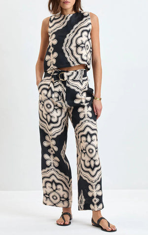 A woman stands wearing the Lemlem Adowa Shell Tank—a sleeveless, asymmetrical top in European flax linen with a bold black and cream abstract floral pattern—paired with wide-leg pants and black sandals.