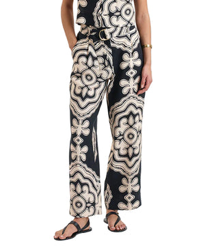 A person wears Lemlem’s Diarra Pant—a relaxed straight-leg style with a white abstract geometric print—paired with a matching European flax linen top, sandals, and a bracelet, standing with one hand in their pocket.