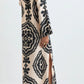 A person wearing the Lemlem Theodora Column Dress—a long, cream-colored linen maxi dress with bold black geometric patterns, long sleeves, and a side slit—paired with flat sandals.