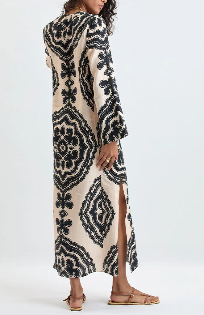 A person wearing the Lemlem Theodora Column Dress—a long, cream-colored linen maxi dress with bold black geometric patterns, long sleeves, and a side slit—paired with flat sandals.