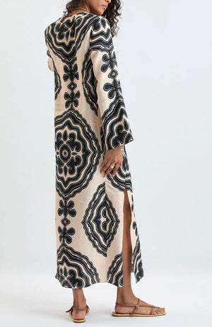 A person wearing the Lemlem Theodora Column Dress—a long, cream-colored linen maxi dress with bold black geometric patterns, long sleeves, and a side slit—paired with flat sandals.