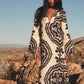 A woman stands outdoors in rocky desert terrain, wearing the Lemlem Theodora Column Dress—a cream-colored linen maxi with bold black geometric patterns and flared sleeves.