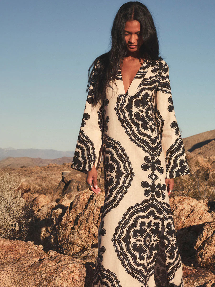 A woman stands outdoors in rocky desert terrain, wearing the Lemlem Theodora Column Dress—a cream-colored linen maxi with bold black geometric patterns and flared sleeves.