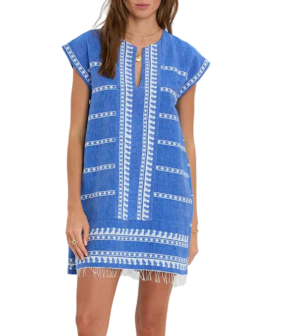 A woman wears the Lemlem Elina Caftan, a blue, knee-length, short-sleeve Ethiopian dress crafted from hand-woven cotton with white embroidery and fringe at the hem.