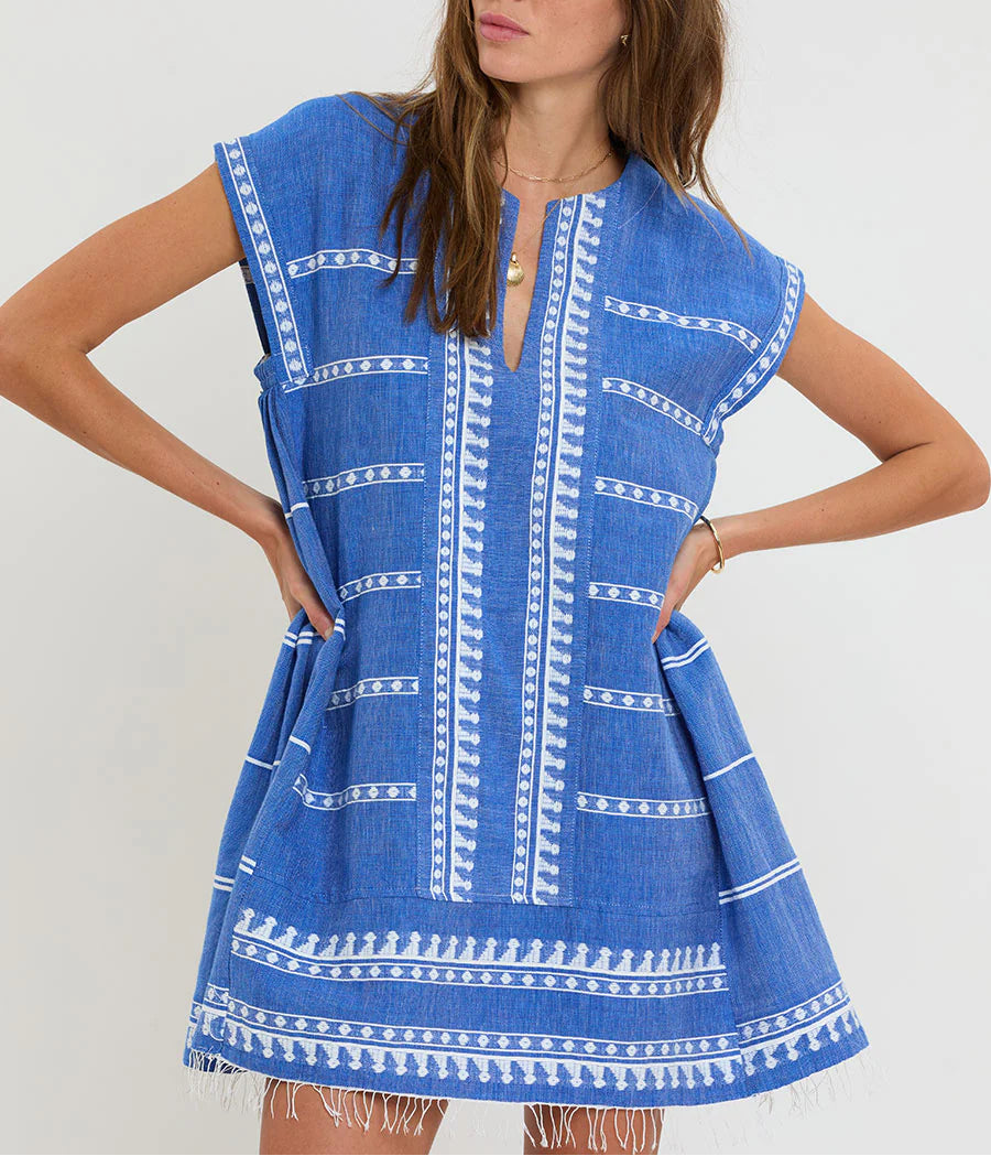 A woman wears the Lemlem Elina Caftan, a short-sleeved blue Ethiopian dress with white embroidery, split neckline, frayed hem, and hand-woven cotton.