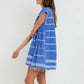 A woman wears the Lemlem Elina Caftan, a loose-fitting, hand-woven cotton blue dress with white stripes and fringe at the hem, offering a modern twist on the classic Ethiopian dress against a simple backdrop.