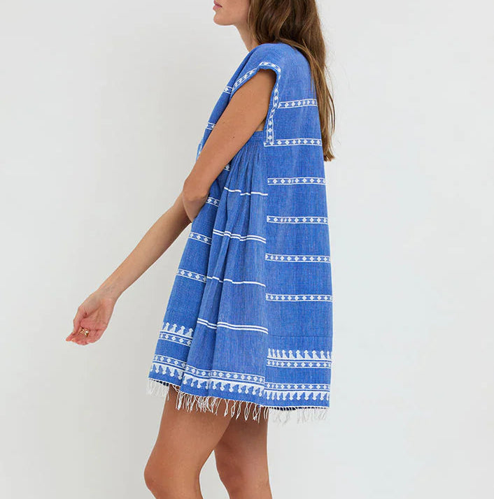 A woman wears the Lemlem Elina Caftan, a loose-fitting, hand-woven cotton blue dress with white stripes and fringe at the hem, offering a modern twist on the classic Ethiopian dress against a simple backdrop.