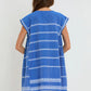A woman with loose brown hair faces away, wearing the Lemlem Elina Caftan—a hand-woven blue cotton cover-up dress with white horizontal patterns, short sleeves, and a fringed hem.