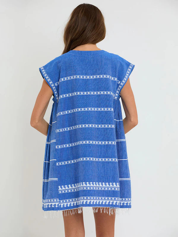 A woman with loose brown hair faces away, wearing the Lemlem Elina Caftan—a hand-woven blue cotton cover-up dress with white horizontal patterns, short sleeves, and a fringed hem.