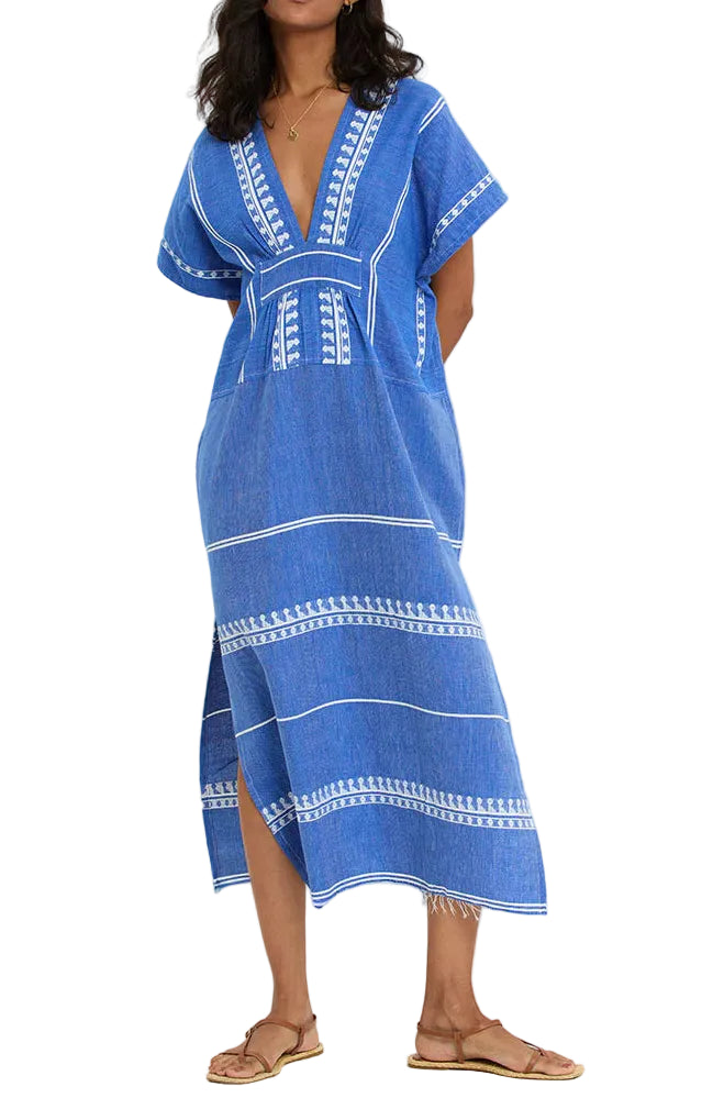 A woman wears the Lemlem Gasira V Neck Caftan, a blue, short-sleeve midi dress with white patterns and stripes, made from hand-woven cotton, paired with flat sandals.
