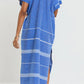 A person wearing the Lemlem Gasira V Neck Caftan, a loose-fitting blue and white patterned dress made from hand-woven cotton, stands facing away to show the back, paired with flat sandals.