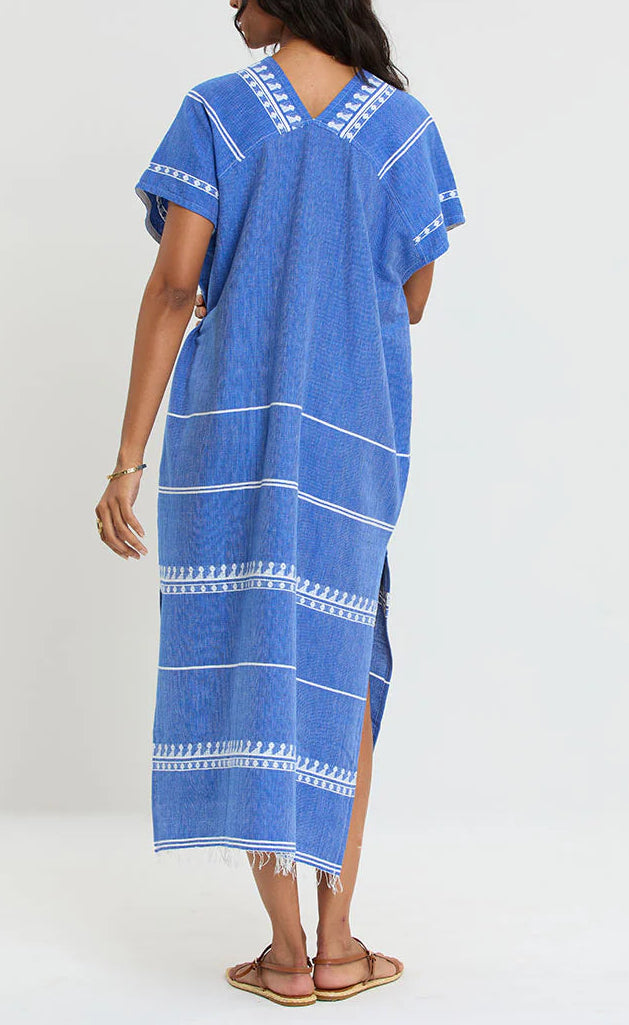 A person wearing the Lemlem Gasira V Neck Caftan, a loose-fitting blue and white patterned dress made from hand-woven cotton, stands facing away to show the back, paired with flat sandals.