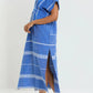 A person in the Lemlem Gasira V Neck Caftan—a blue and white hand-woven cotton dress from Ethiopia with a side slit and frayed hem—pairs it with brown sandals against a plain background.
