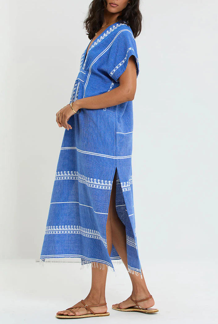 A person in the Lemlem Gasira V Neck Caftan—a blue and white hand-woven cotton dress from Ethiopia with a side slit and frayed hem—pairs it with brown sandals against a plain background.