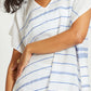 A person wears the Lemlem Dalila Caftan—a hand-woven white piece with blue horizontal stitches, short sleeves, and a V-neck—resting their hand on their torso to show a gold ring.