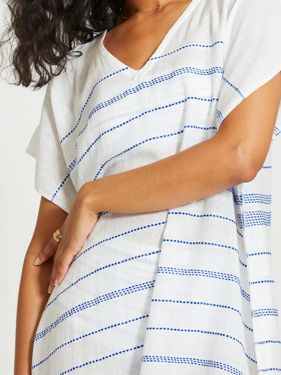A person wears the Lemlem Dalila Caftan—a hand-woven white piece with blue horizontal stitches, short sleeves, and a V-neck—resting their hand on their torso to show a gold ring.