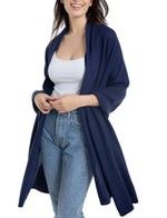 A woman, smiling with arms slightly bent, is elegantly draped in the Alashan Cashmere Cashmere Luxe Travel Wrap over a white tank top, blue open-front cardigan, and light blue jeans.
