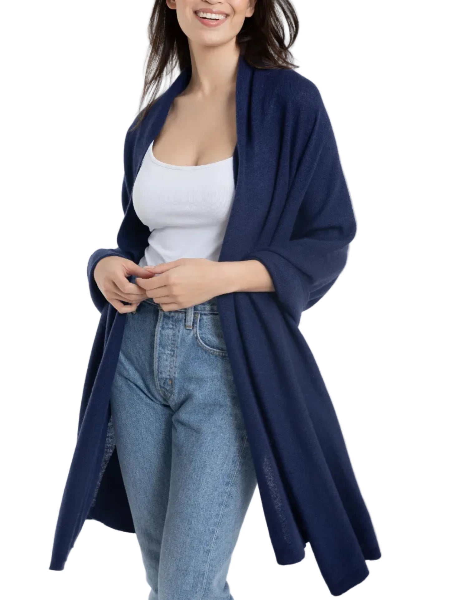 A woman, smiling with arms slightly bent, is elegantly draped in the Alashan Cashmere Cashmere Luxe Travel Wrap over a white tank top, blue open-front cardigan, and light blue jeans.