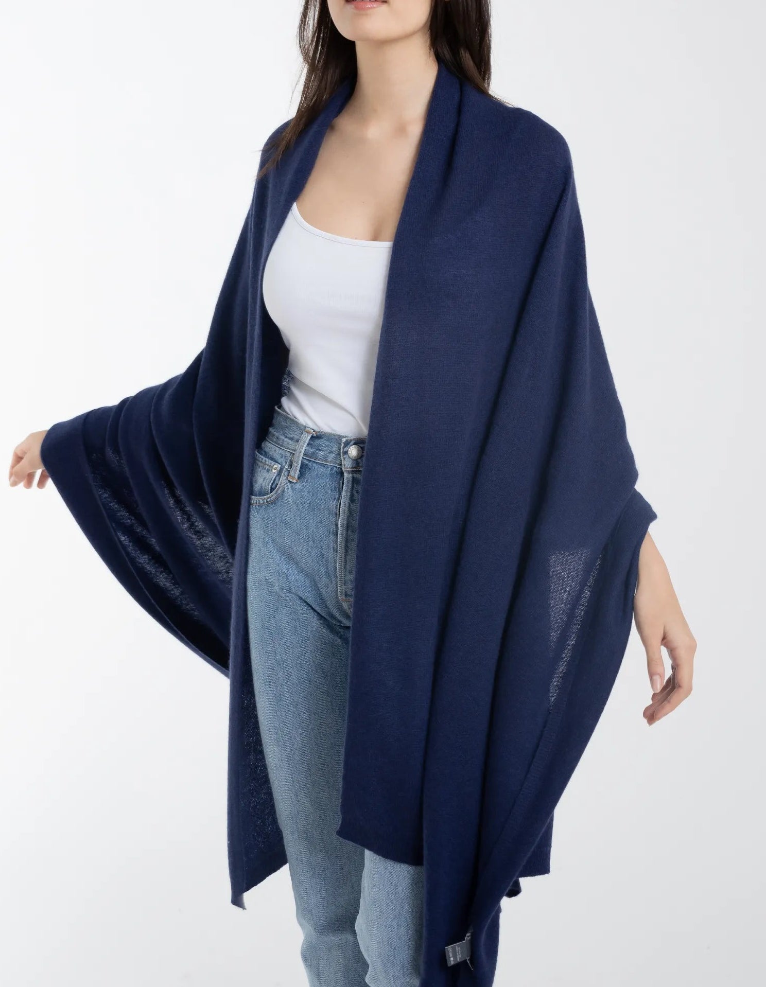 A woman stands against a plain white background wearing a white top, blue jeans, and the Alashan Cashmere Luxe Travel Wrap in navy blue.
