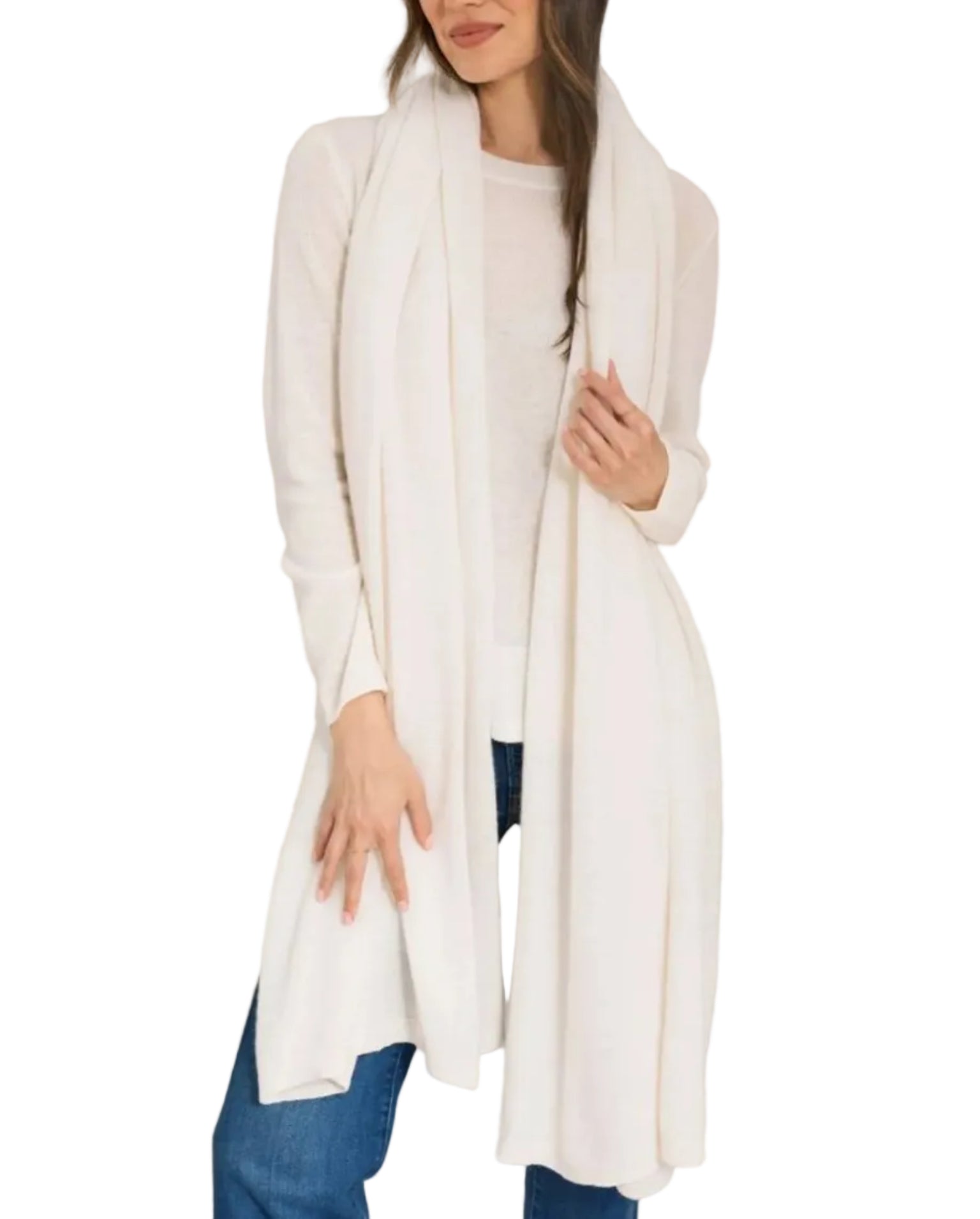 A woman stands against a white background wearing the Alashan Cashmere Cashmere Luxe Travel Wrap—a long, light cream, luxurious scarf—draped over a matching sweater and blue jeans.